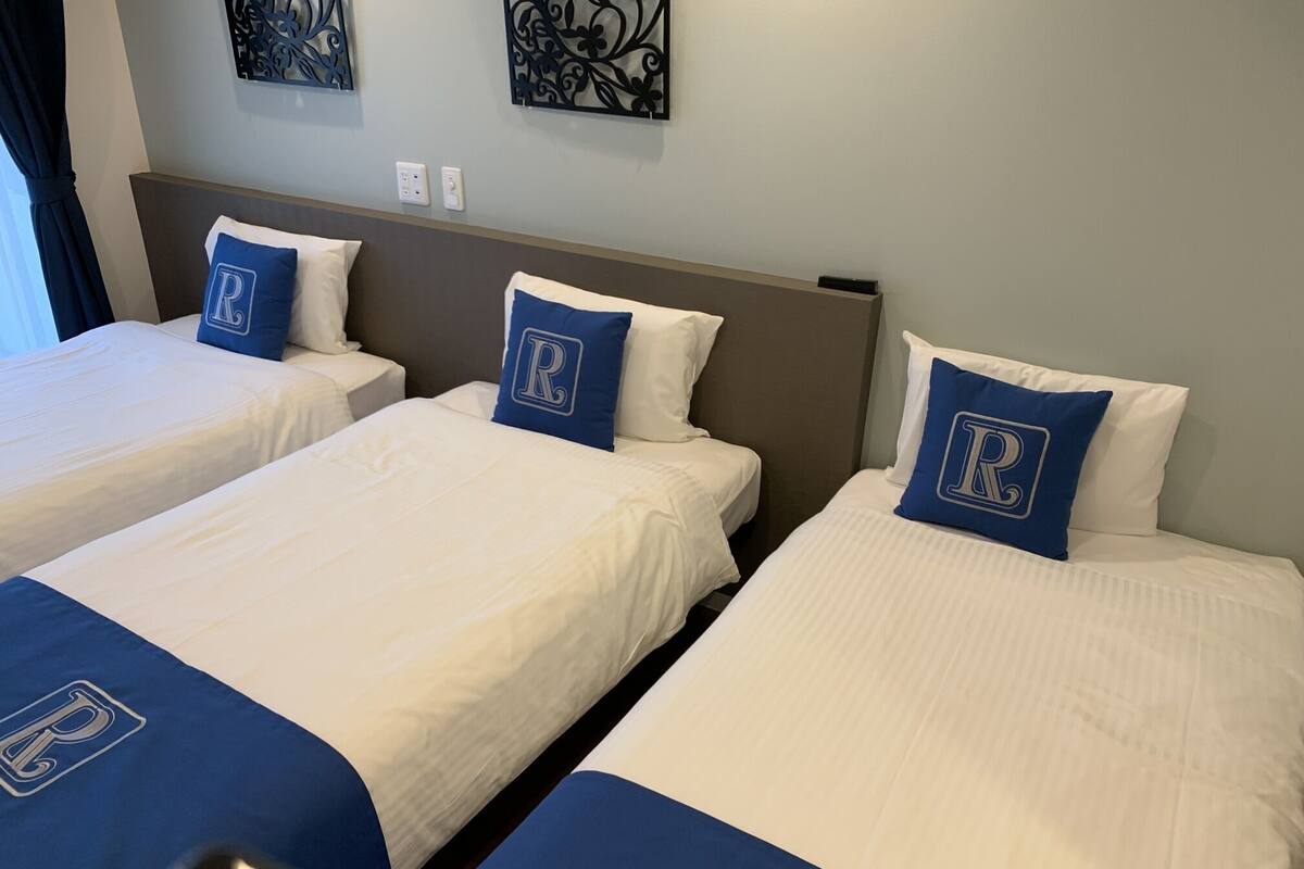 Three twin beds with white bedding and blue headboards, each with a pillow featuring a large 'R'.