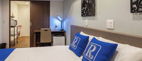 A hotel room with a bed, a desk, and a chair. There is a wall decoration above the bed and a fire extinguisher on the wall.
