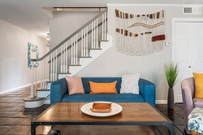 Un salon avec un canapé bleu, une table basse en bois, un escalier et une décoration murale en macramé.