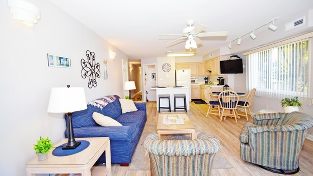A modern living room with a blue sofa, a coffee table, and striped armchairs. There is a kitchen area with wooden cabinets and a dining table. - Coral Ridge 1-Oceanside 78th St, Free WiFi, W/D, AC (Ocean City)