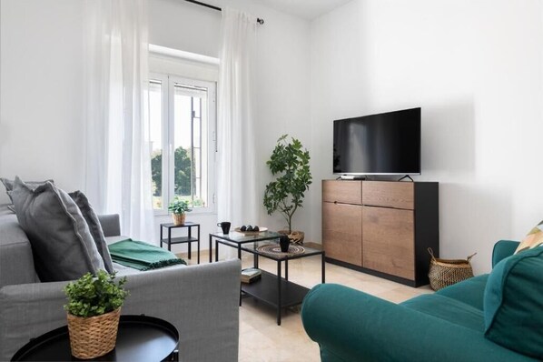 A modern living room with a grey sofa, a teal sofa, a coffee table, a TV on a wooden cabinet, and a window with curtains.