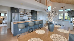 A modern kitchen with a central island, bar stools, and a dining table with placemats. - Experience ocean views at Pelican Brief, a spacious 4-bedroom house in Oak Island! (Oak Island)