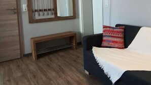 Um quarto com piso de madeira, um sofá escuro com almofada estampada, um cobertor branco, um banco de madeira e um espelho com moldura de madeira fixado na parede.