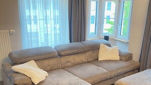 Uma sala de estar moderna com um amplo sofá em formato de L e um banquinho combinando, um radiador e vista para o exterior por grandes janelas.