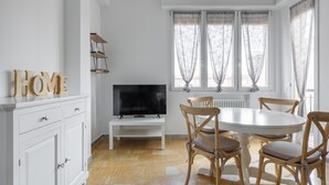 A dining area with a round table, four wooden chairs, a television on a stand, and a white cabinet with drawers.
