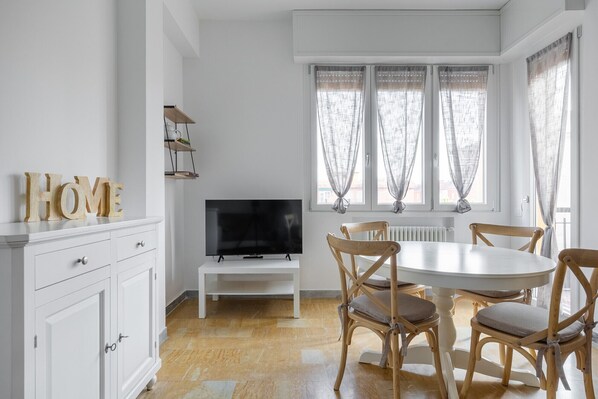 A dining area with a round table, four wooden chairs, a television on a stand, and a white cabinet with drawers.