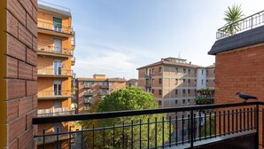 A balcony with a view of multi-story buildings, a bird on the railing, and a tree in the foreground.