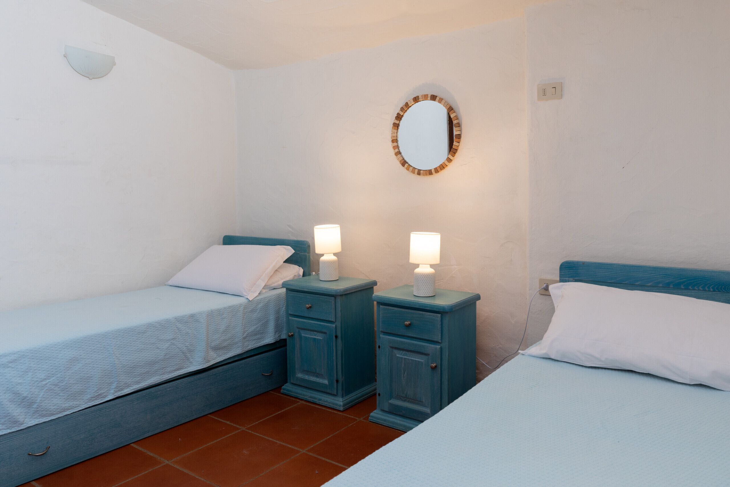 Quarto com duas camas, cada uma com travesseiro branco e cabeceira azul. Há duas mesas de cabeceira com luminárias, e um espelho na parede.