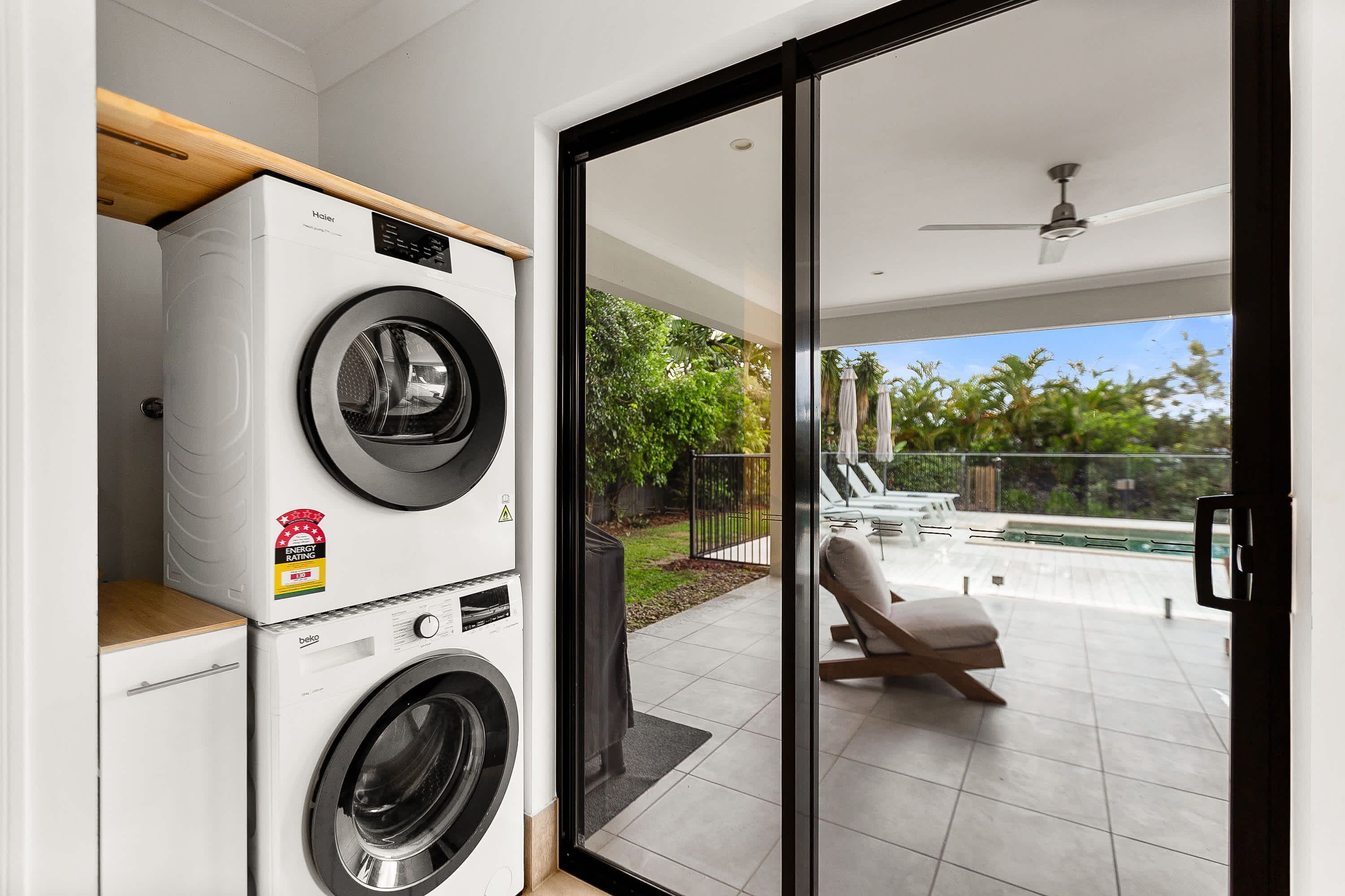 A laundry area with a front-loading washer and dryer, a wooden shelf, and a sliding glass door leading to an outdoor patio with a chair and umbrella.