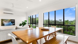 A modern dining area with a wooden table, white chairs, and large windows offering a view of greenery. - Cove Retreat - Kid Pet and Pool Paradise (Palm Cove)