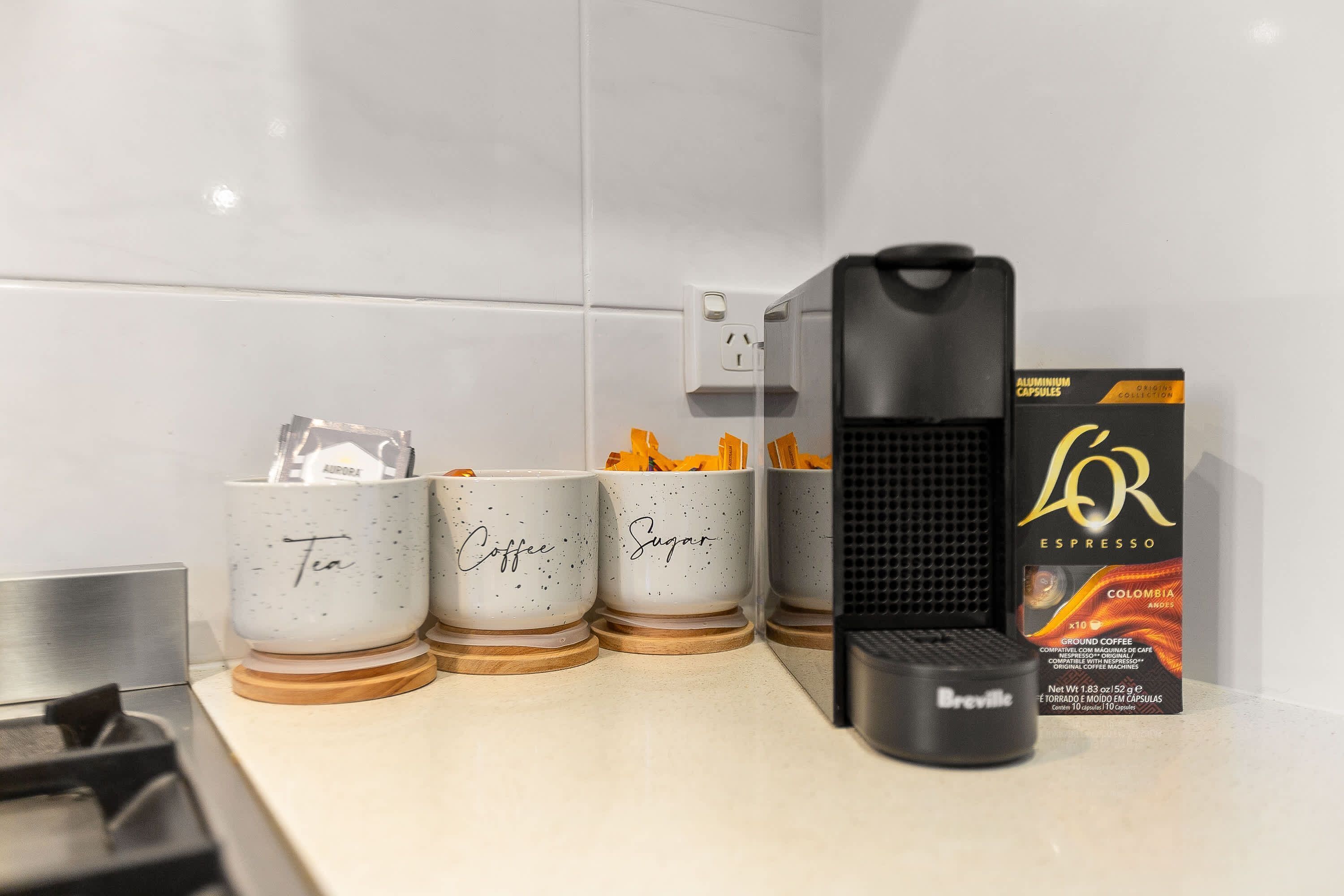 A kitchen counter with three white mugs labeled "Tea," "Coffee," and "Sugar," a coffee maker, and a box of L'Or espresso coffee capsules.