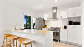 A modern kitchen with white cabinetry, a large island with stools, stainless steel appliances, and a view of greenery outside. - Cove Retreat - Kid Pet and Pool Paradise (Palm Cove)