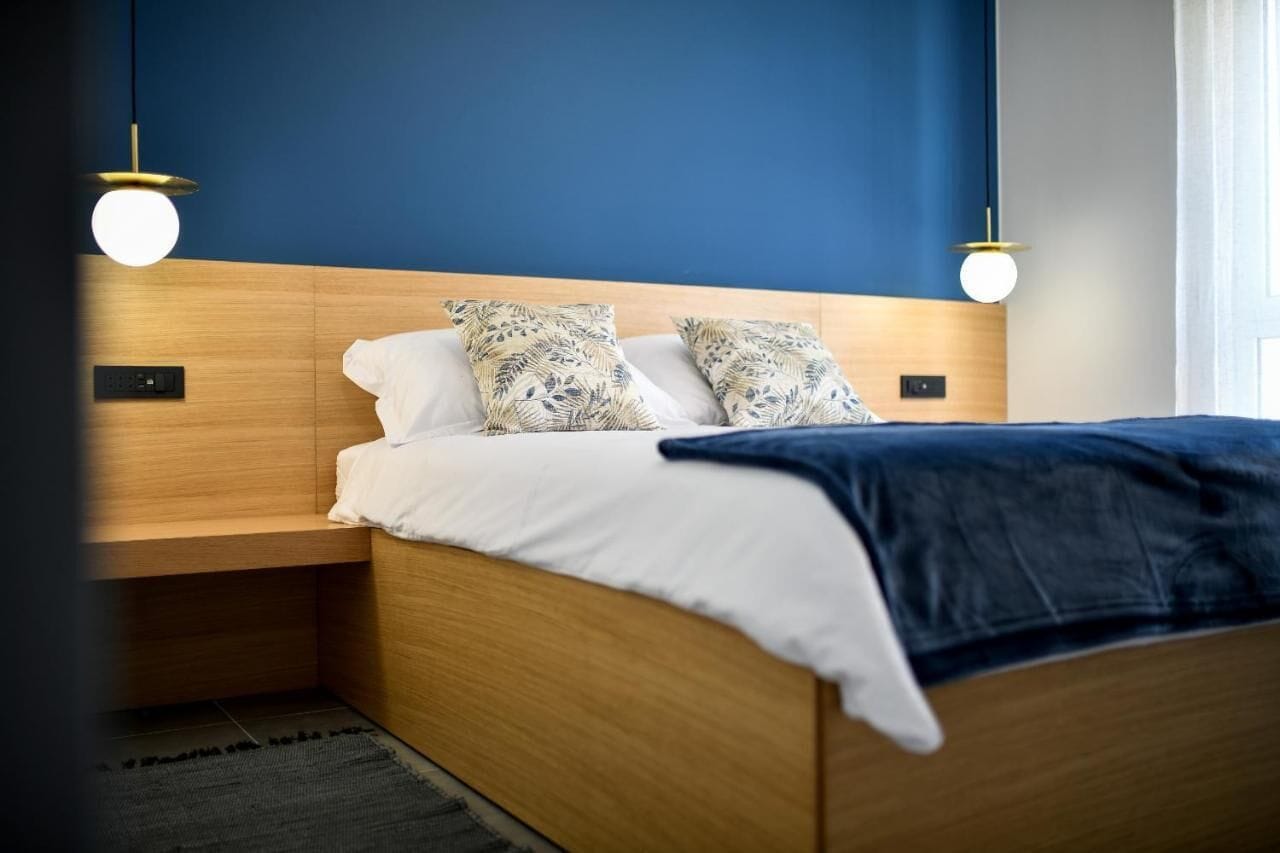 A modern bedroom with a wooden headboard, white bedding, patterned pillows, and two pendant lights.