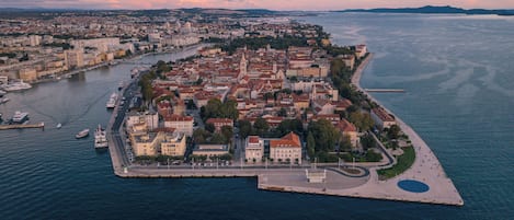 An aerial view of a coastal city with a prominent peninsula, featuring a mix of historical and modern architecture, a marina with boats, and a large body of water.