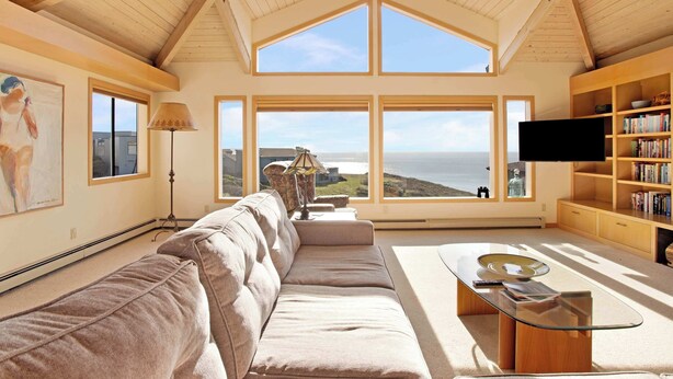A spacious living room with a large window, a sofa, a coffee table, a bookshelf, and a flat-screen TV. - Amazing Oceanview's - Dog-Friendly 4BR with Sauna (Bodega Bay)