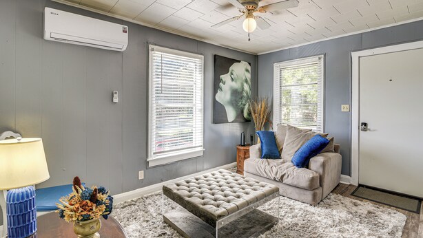 A living room with a tufted ottoman, a sofa, a lamp, and a wall-mounted air conditioner. - Near Sabine Lake & Refineries: Pet-friendly Apt (Port Arthur)