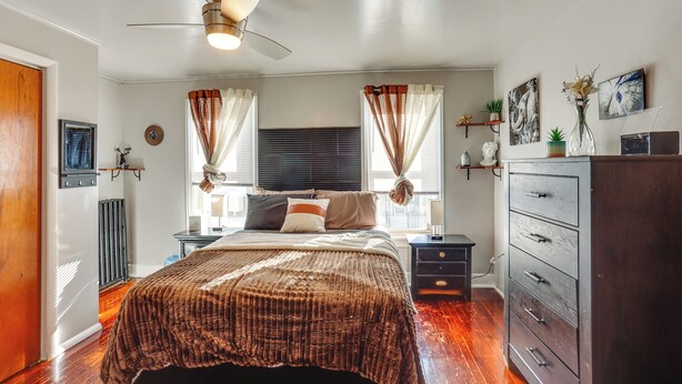 Une chambre à coucher comprenant un lit, une commode, une table de chevet, un ventilateur de plafond et une fenêtre avec des rideaux.