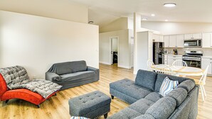 Een moderne woonkamer met een grijze bank, een rode fauteuil en een poef, een eethoek met een houten tafel en stoelen, en een keuken met witte kasten en apparatuur.