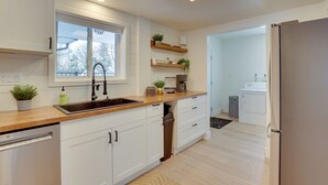 Una cocina moderna con armarios blancos, encimera de madera, un lavaplatos integrado y un refrigerador. Hay plantas en macetas sobre la encimera y estantes encima del lavaplatos.