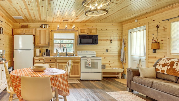 A cozy cabin interior with wooden walls, a dining area with a checkered tablecloth, a kitchenette with modern appliances, and a comfortable seating area with a leather couch. - On 40 Acres: Charming Cabin in Zanesville! (Zanesville)