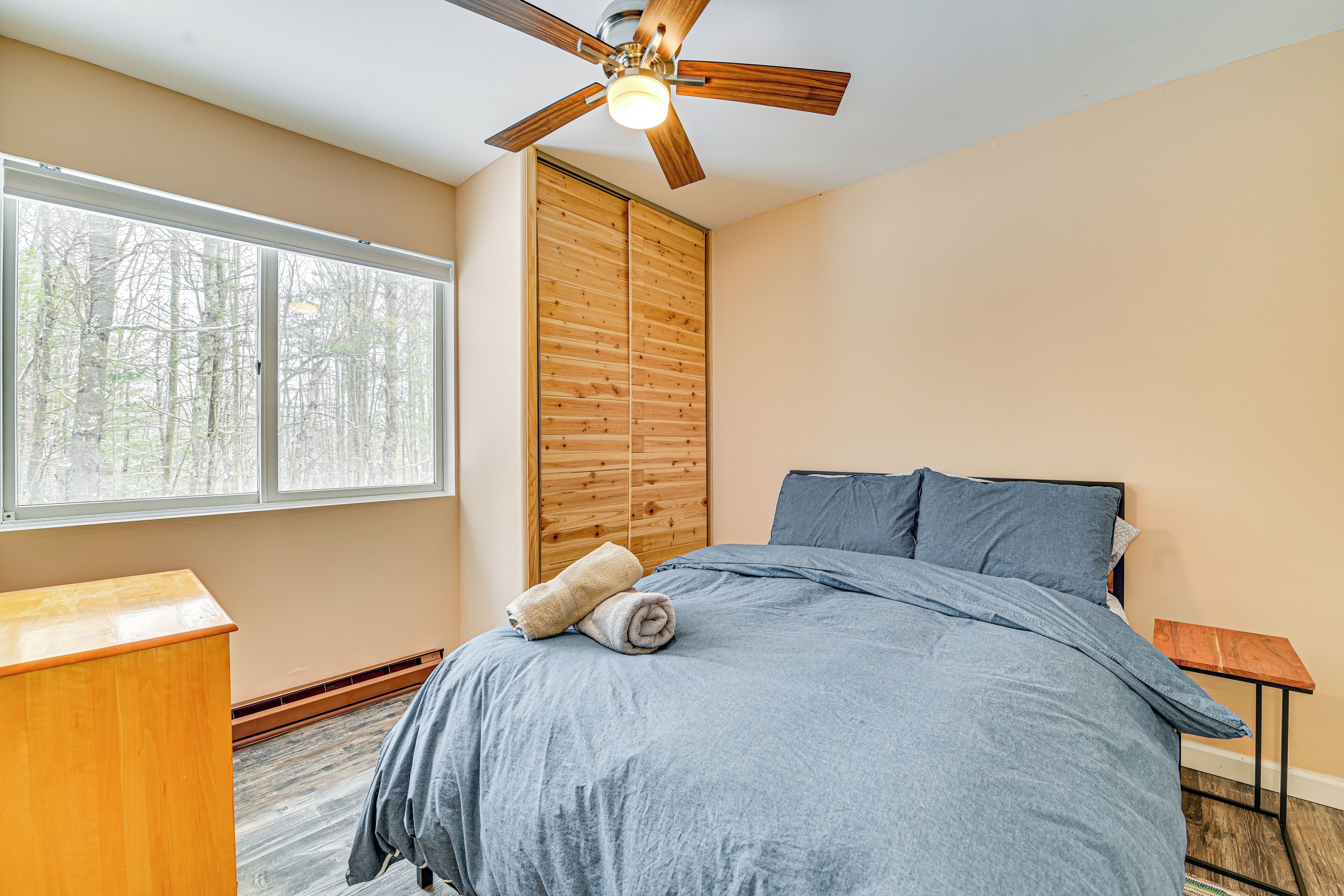 A bedroom with a bed, a wooden wardrobe, a small wooden table, and a ceiling fan.