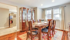 Un comedor con mesa de madera, sillas y un tapete rojo con estampado. Se observa un armario con puertas de cristal y un refrigerador al fondo.