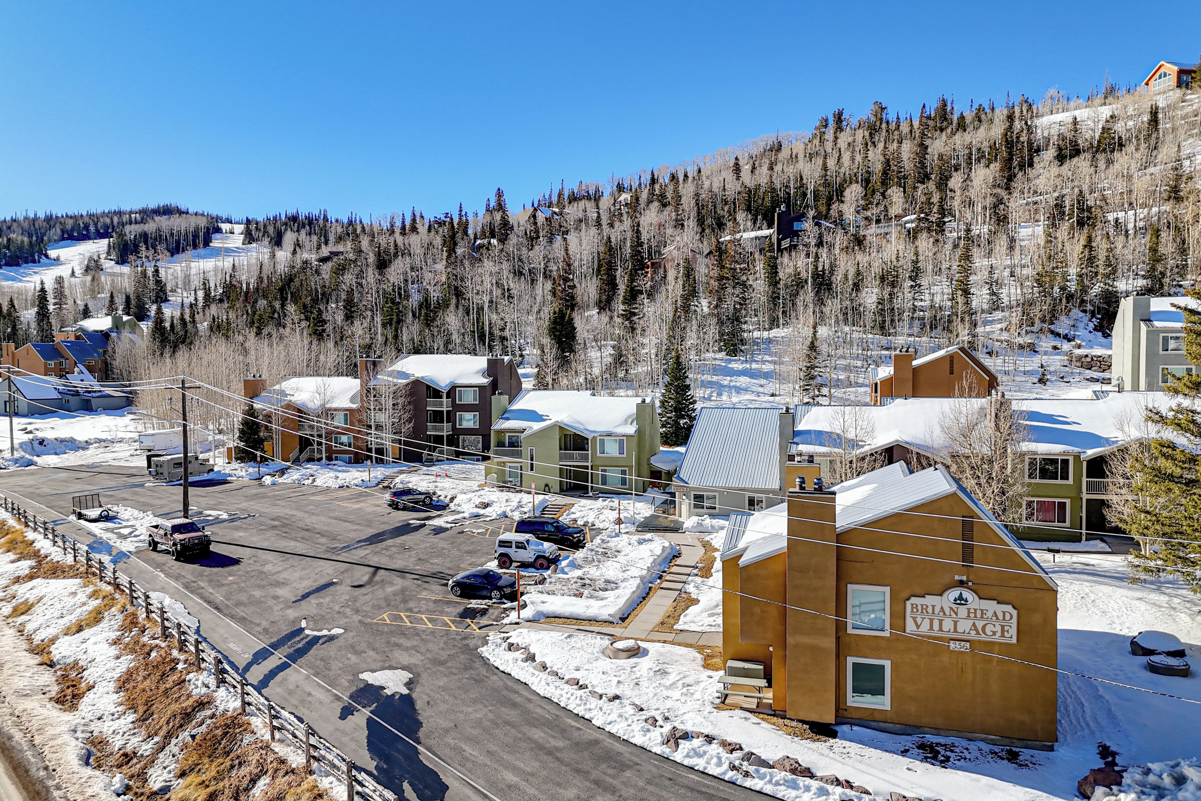 A residential area with a Brian Head Village sign, snow-covered houses, and a parking lot with several cars.