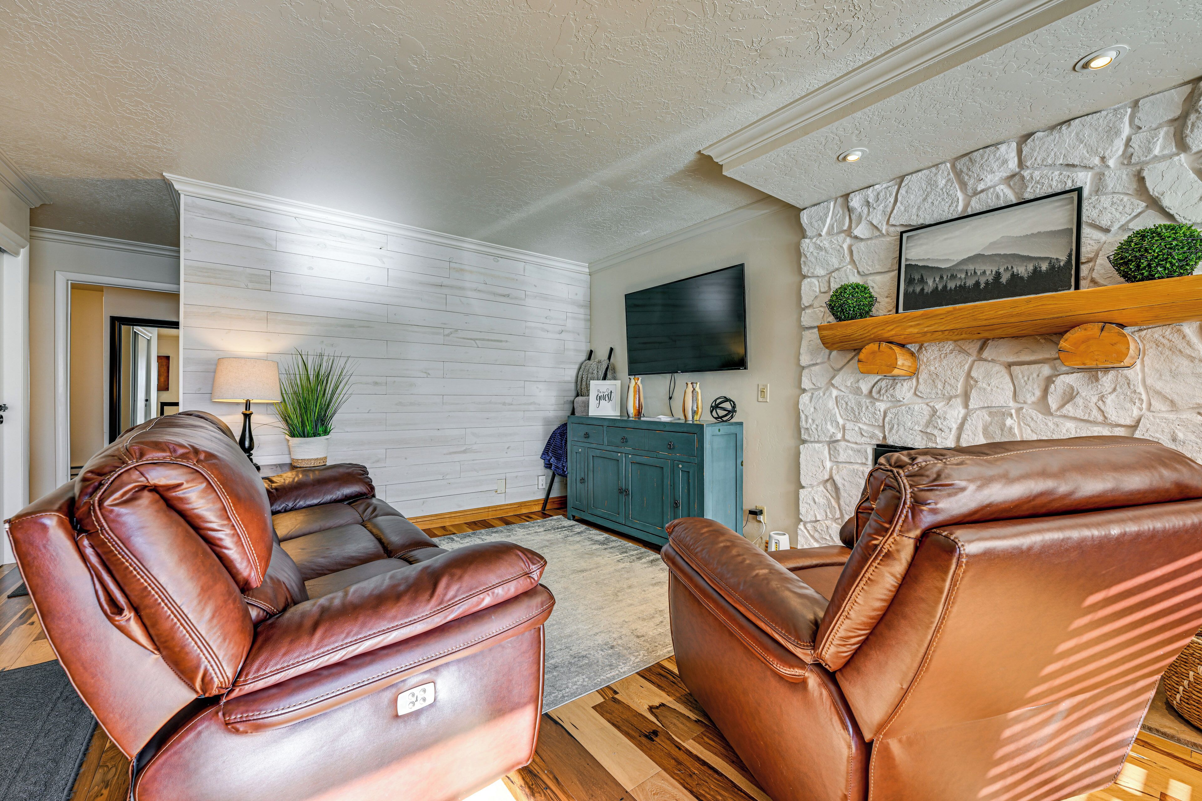 A living room with leather sofas, a stone wall, a mounted TV, and a wooden shelf with decorative items.