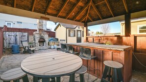 Un patio al aire libre con una mesa redonda y taburetes, una chimenea de piedra y una valla de madera.