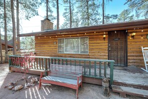 Une cabane rustique en rondins de bois, dotée d’une terrasse en bois, d’un banc et d’une porte sur laquelle est accrochée une enseigne.