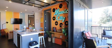 Una cocina moderna con una isla central, un área de comedor con mobiliario de mimbre y una sala de estar con un cartel que dice 'Music City'.