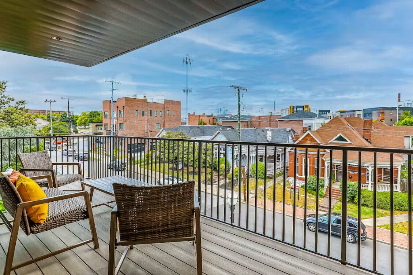 Un balcón con mobiliario de mimbre y vistas a un barrio residencial con casas de ladrillo y una calle.