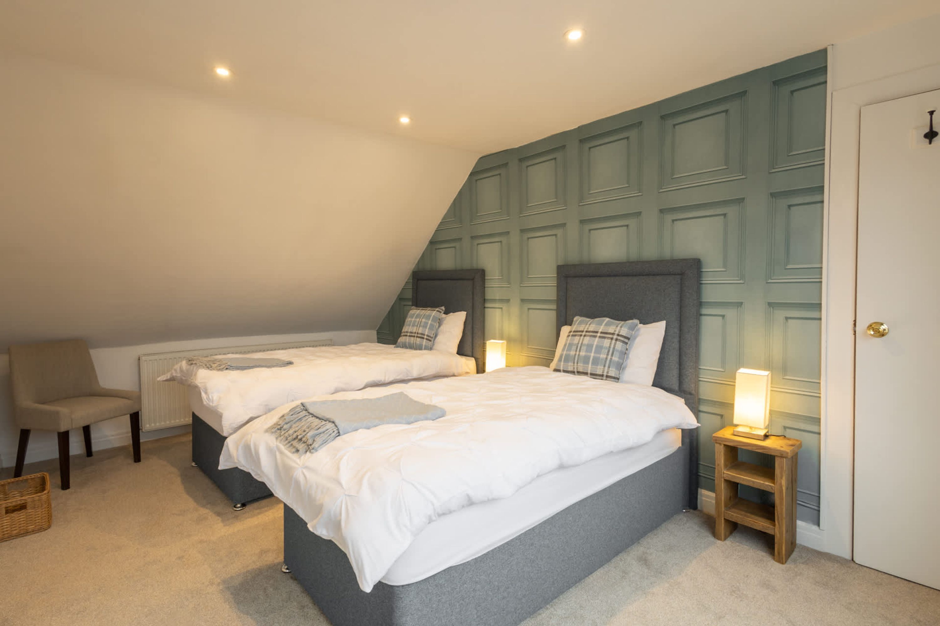A bedroom with two beds, a chair, a bedside table with a lamp, and a door.