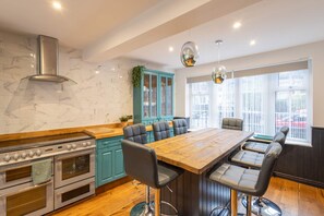 A modern kitchen with a large island, built-in oven, teal cabinetry, and a view of the city through large windows. - Host Stay The Coachman s House (Harrogate)