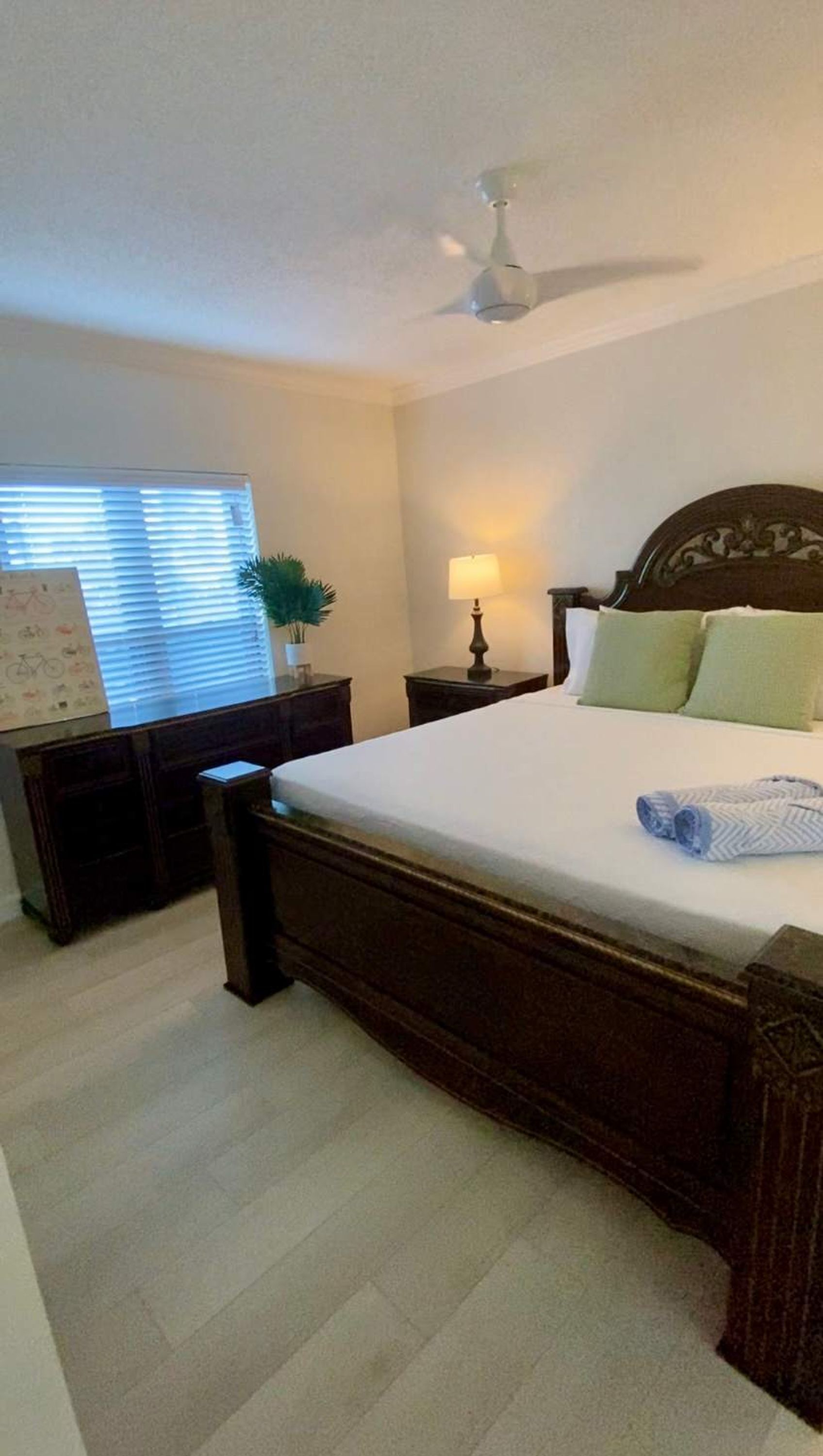 A bedroom with a large bed, a nightstand, a lamp, a ceiling fan, and a window with blinds.