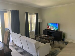 A living room with a sofa, a television, a coffee table, and a window with curtains. - Oceanfront Condo on Seven Mile Beach (Seven Mile Beach)