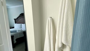 Un baño con toallas blancas colgadas en un perchero, una cortina de ducha azul y una puerta visible que conduce a otra habitación con una cama y una mesita de noche.