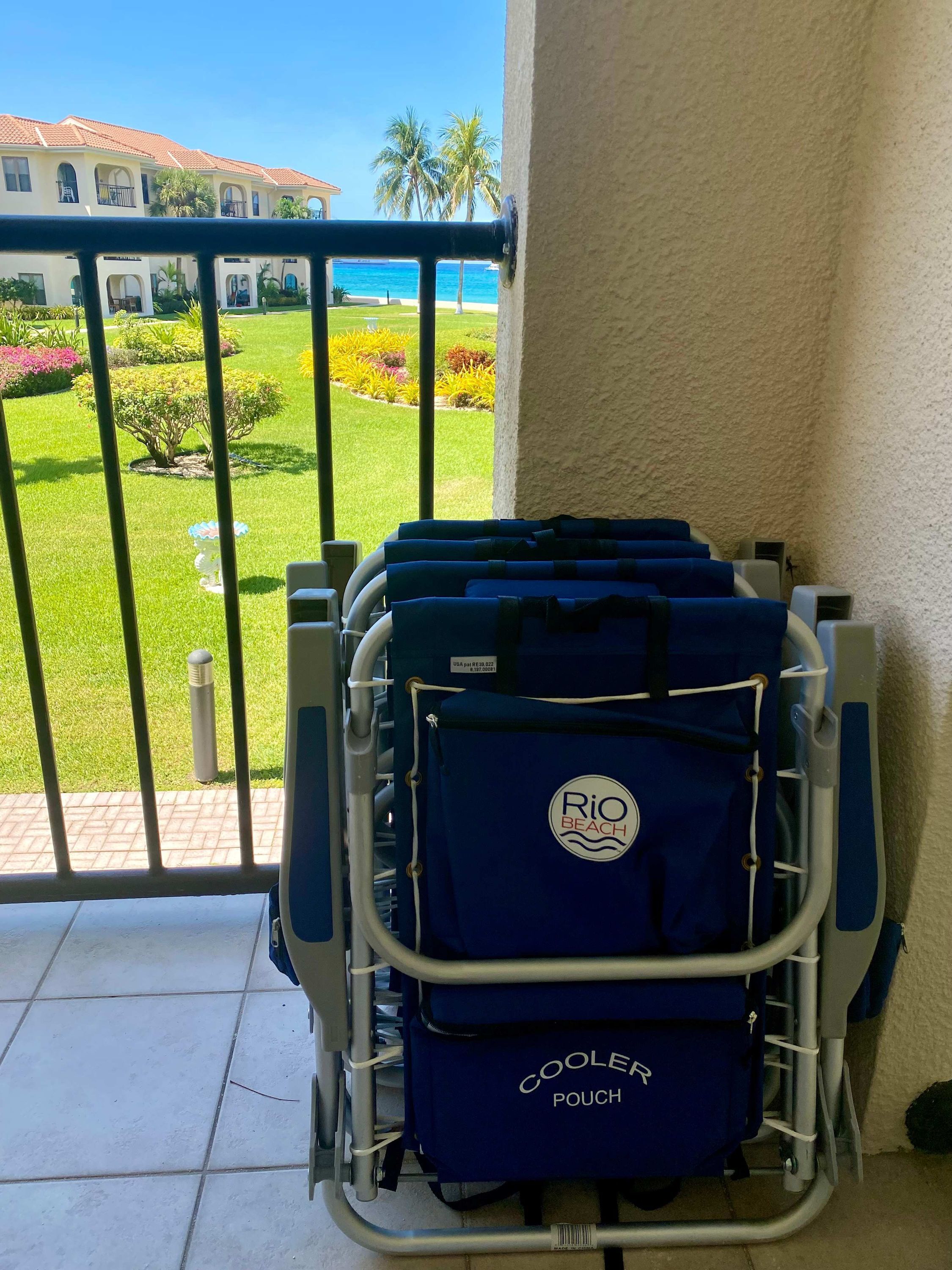 A luggage cart with a 'Rio Beach' pouch on a balcony overlooking a landscaped garden and a building with a tiled roof.