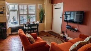 A living room with orange sofas, a dining table with chairs, a TV mounted on the wall, and a large window with blinds. - Host Stay The Bothy (Lyme Regis)