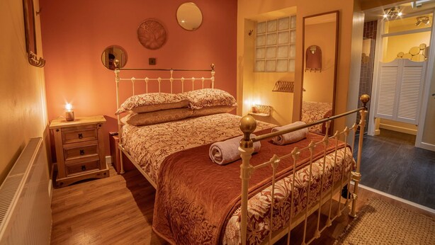 A bedroom with a bed, bedside tables, a mirror, and a window with blinds. - Host Stay The Bothy (Lyme Regis)