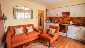 A compact living space with a sofa and armchair, a kitchen with white cabinets and wooden backsplash, and a mirror on the wall. - Host Stay The Bothy (Lyme Regis)