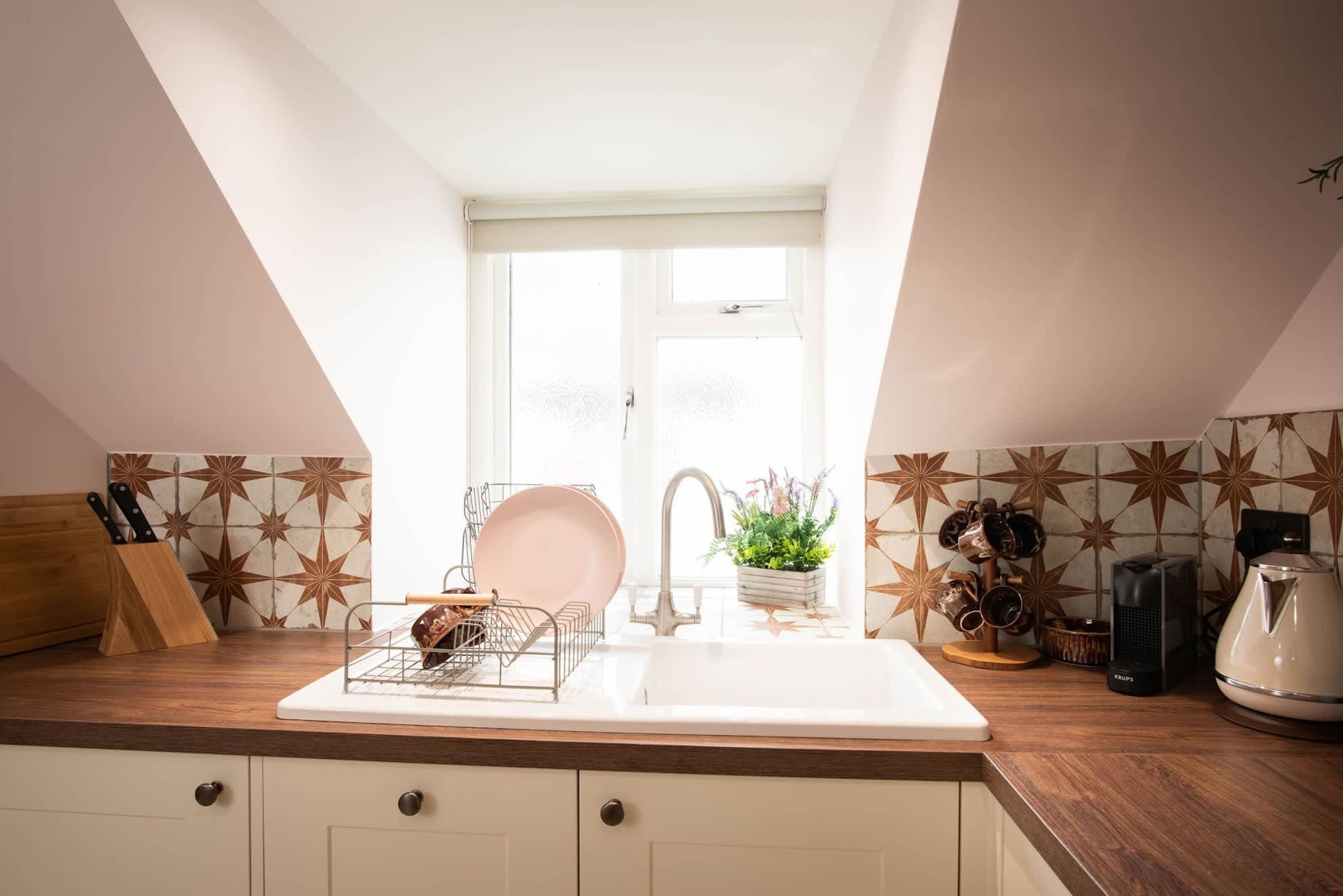 Una cocina con mostrador blanco, un lavamanos y una ventana encima. Hay utensilios de cocina y una tetera en el mostrador.