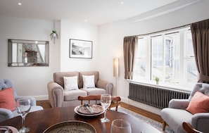 A modern living room with a sofa set, a dining table with a plate and wine glasses, a large mirror, and a window with curtains.