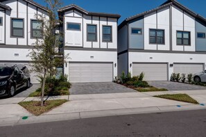 A row of modern two-story townhouses with white exteriors and grey trim, featuring front garages and a small tree in the driveway. - Amazing 5BD Family Townhouse at Paradiso Grande (Orlando)
