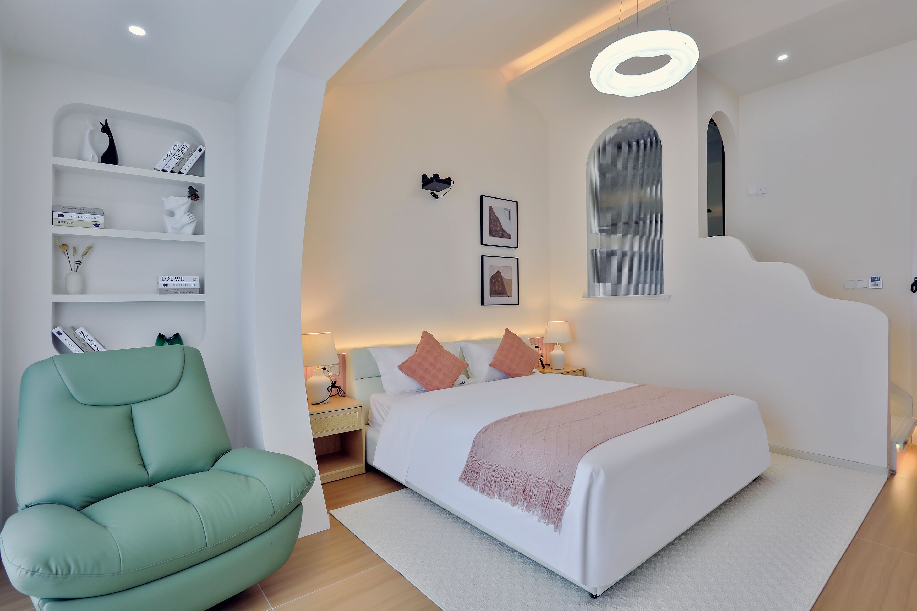 Un dormitorio moderno con un sillón verde, una cama grande con una manta rosa, una mesita de noche blanca y un proyector montado en la pared.