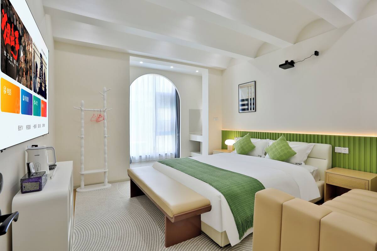 A modern hotel room with a large bed, a bench, a desk with a lamp, and a wall-mounted TV.