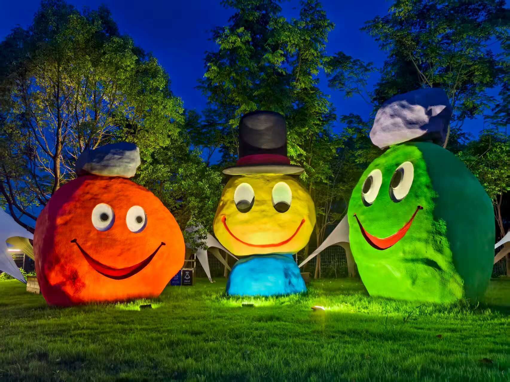 Three large, colorful, spherical sculptures with faces, each wearing different hats, set on grass with trees in the background.