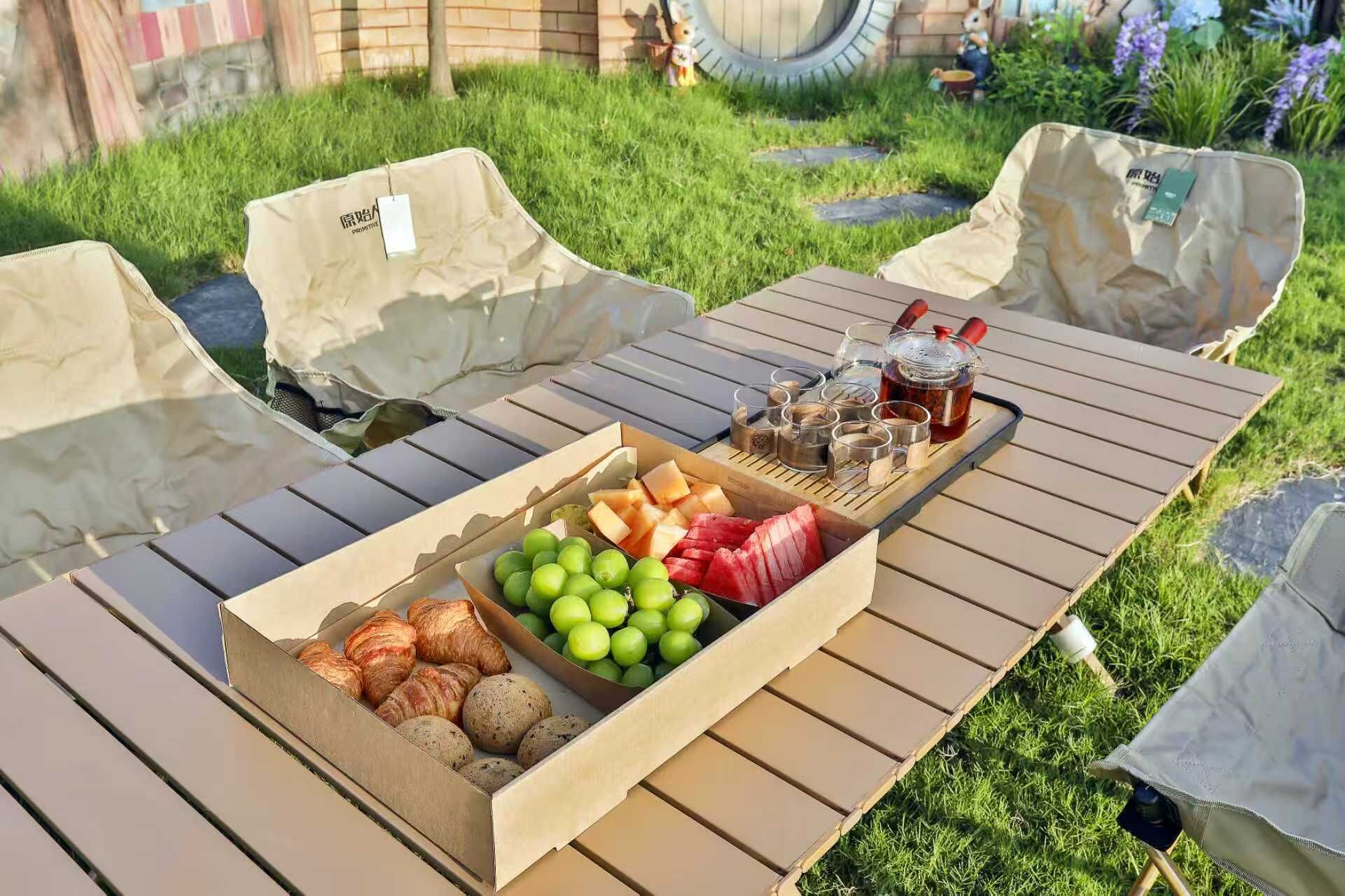 Un espace de restauration en plein air avec une table, des chaises et une boîte contenant des aliments, notamment des fruits, des pâtisseries et des condiments.