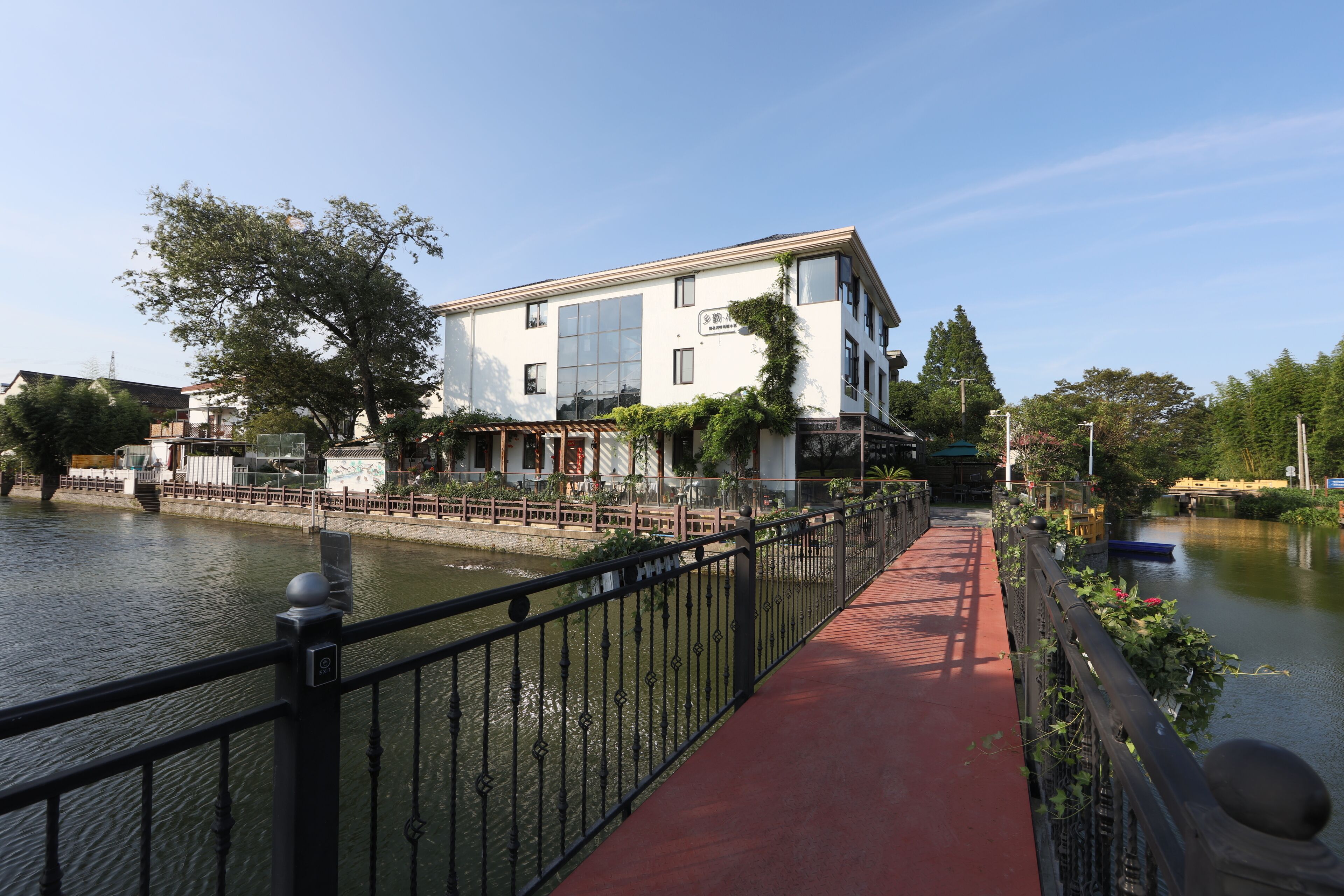 A riverside walkway with a red path, a white building with large windows, and a bridge in the background.