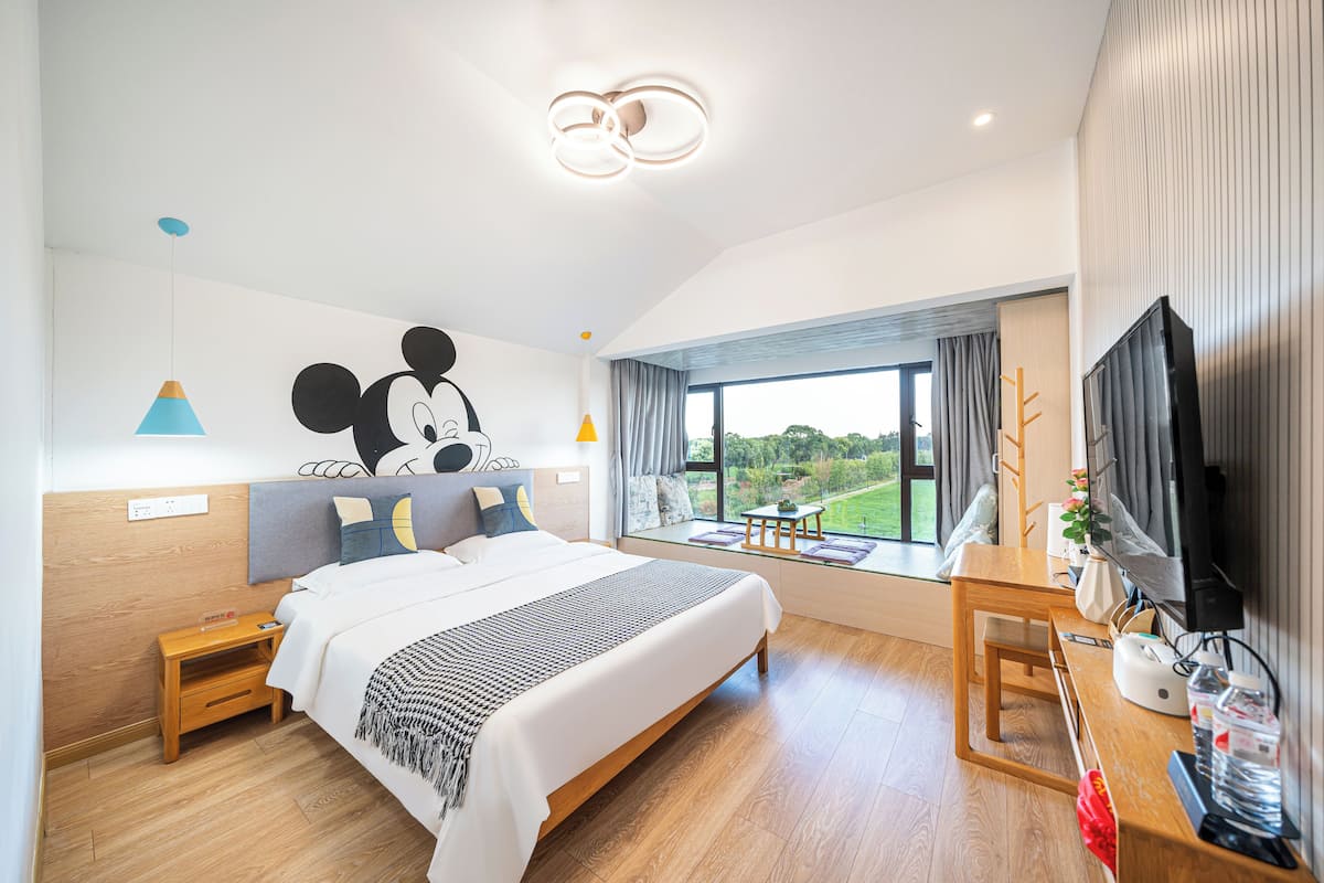 A modern hotel room with a large bed, a desk with a TV, a window with a view of greenery, and a Mickey Mouse mural on the wall.